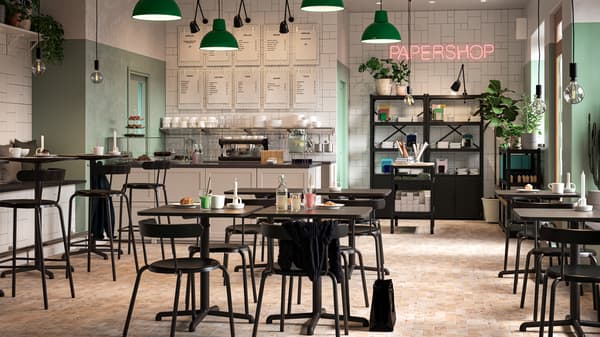Kafe yang mempunyai meja dan kerusi berwarna hitam, dinding berwarna hijau dan putih, kedai kertas, lampu pendan berwarna hijau dan tumbuh-tumbuhan.