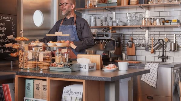 カフェのカウンターにパンとコーヒーカップが並び、奥にエプロン姿の店員と調理器具が見える。