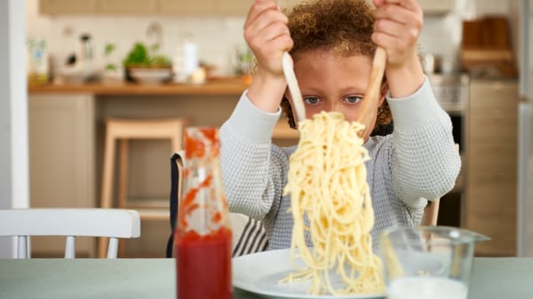 Kahte puidust söögiriista hoides asetab halli kampsunit kandev laps keedetud spagetid valgele IKEA 365+ taldrikule.