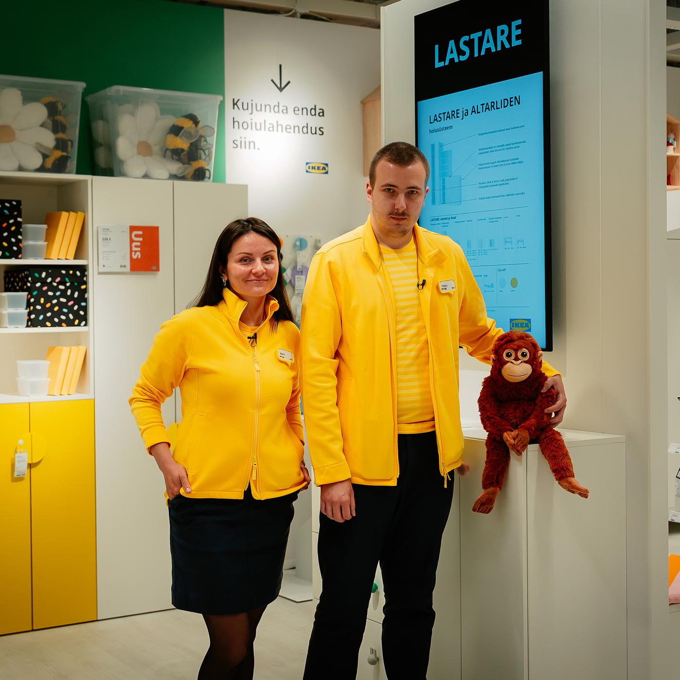 Kaks IKEA töötajat kollastes vormiriietes seisavad Tallinna poe lasteosakonnas riiulite ja väljapanekute kõrval; üks hoiab kätt mänguasjast ahvi ümber, taustal on laste mööbel ja ekraan tekstiga „LASTARE“.