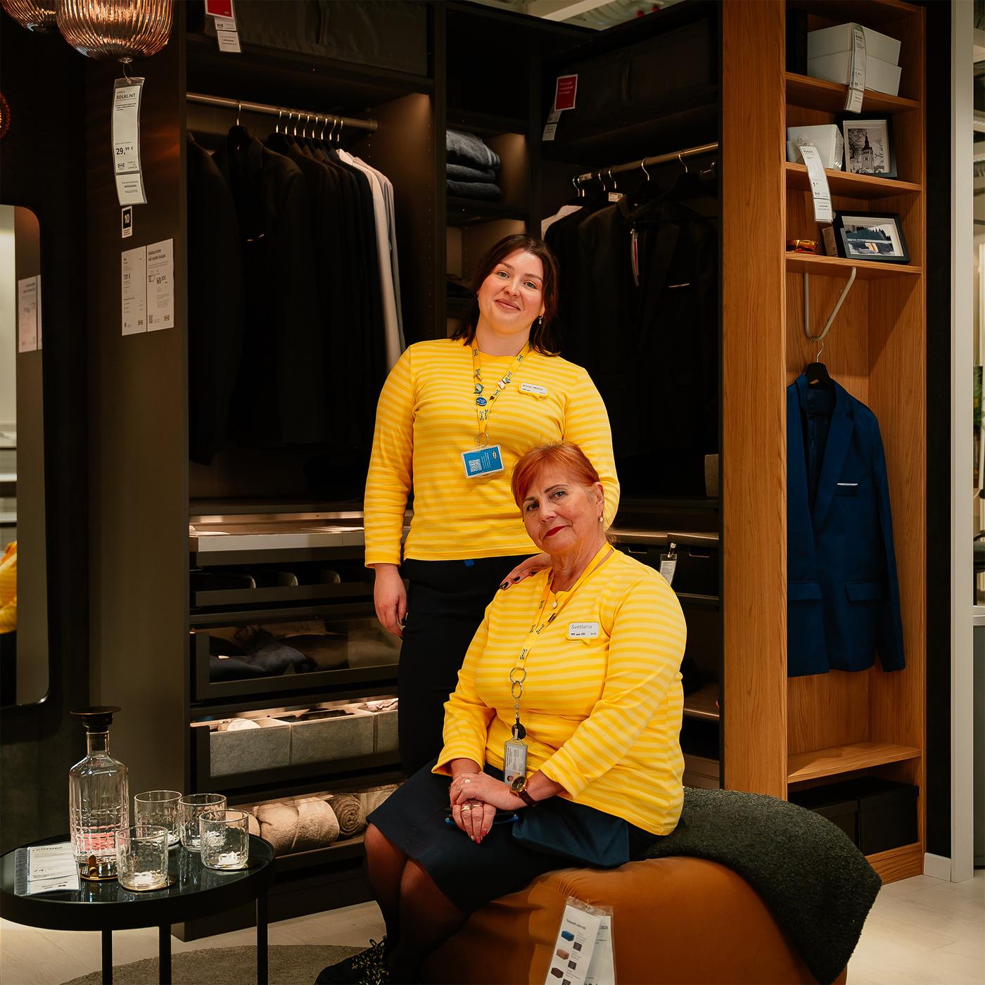 Kaks IKEA töötajat PAX garderoobisüsteemi väljapaneku ees. Üks istub tumedal tumbal, teine seisab avatud garderoobi kõrval, kus on näha riiulid, sahtlid ja riputuslahendused.