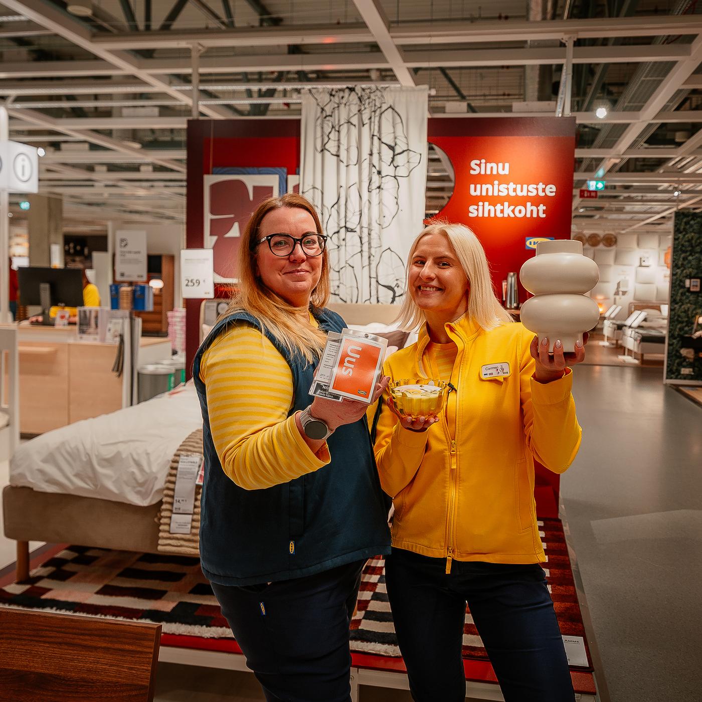 Kaks sinikollastes vormirõivastes rõõmsat IKEA töötajat IKEA Tallinna poe näidistesaalis koos erinevate uudistoodetega, nagu vaas ja küünal.