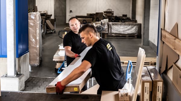 Kaksi mustiin t-paitoihin ja punaisiin työhansikkaisiin pukeutunutta miestä nostaa litteitä IKEA-pakkauksia kuorma-auton perälle.