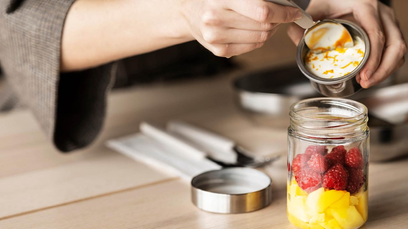 Kanalat tartó kezet látunk, amint egy kisebb fémdobozból joghurtot kanalaz egy DAGKLAR gyümölcstároló dobozba.