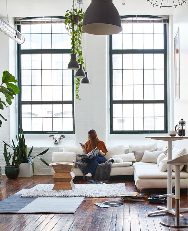 Kara sitting on a white sofa in a living room with high ceilings, large windows and wooden floors.