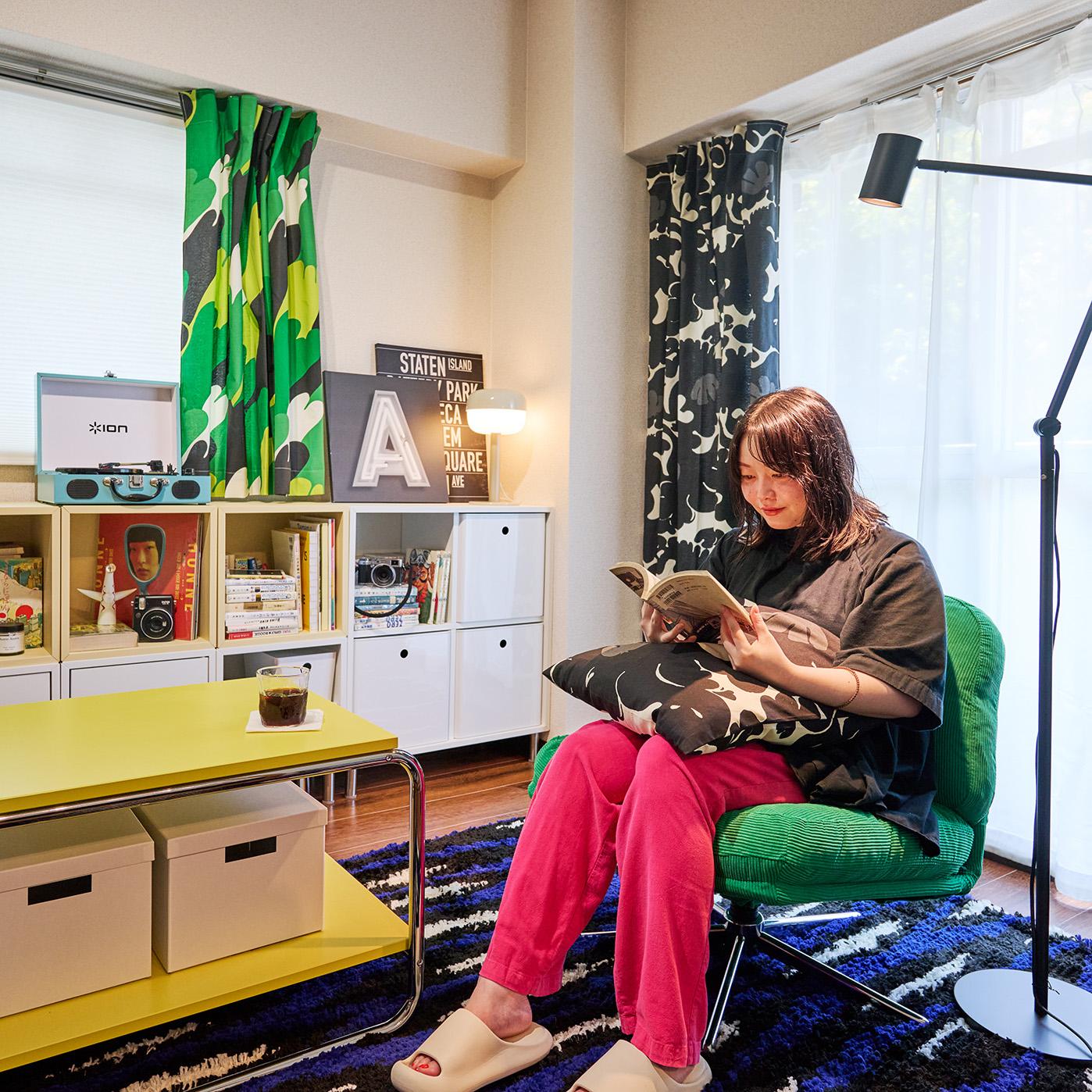 カラフルなカーテンと棚のある居心地の良い部屋で、緑の椅子に座って本を読む人物。