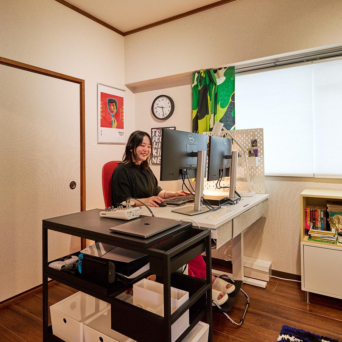 カラフルなカーテンと整理された棚のあるホームオフィスで、デュアルモニターを使って作業する人物。