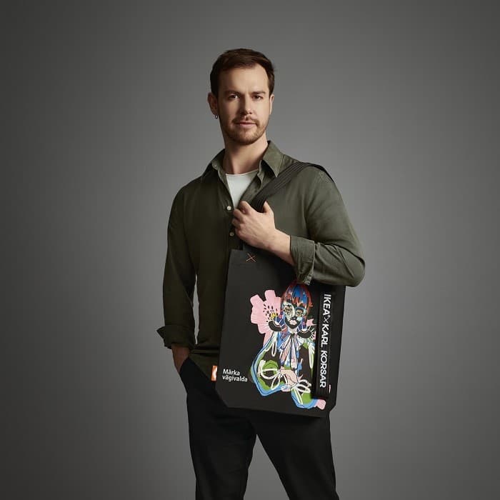Karl Korsar, dressed in an olive-green shirt and black trousers, stands against a grey background with the IKEA “Notice Violence” bag he designed on his shoulder.
