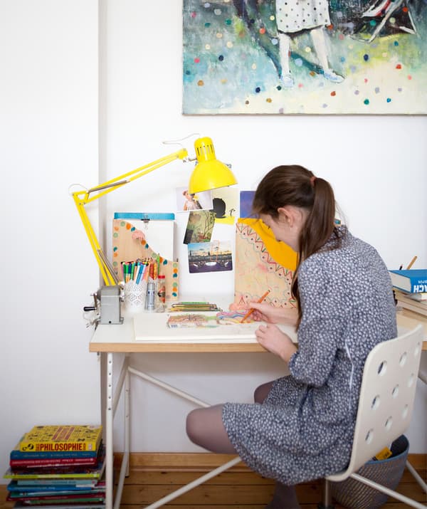 Karlotta dessinant à son bureau sur lequel sont posés une lampe de travail jaune et des range-revues.