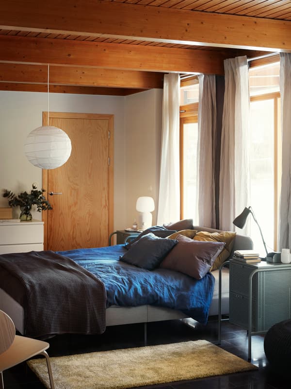 Kárpitozott SLATTUM ágy áll a napfényes és élénk színű, modern hálószobában, az ágyon kék takaró. 