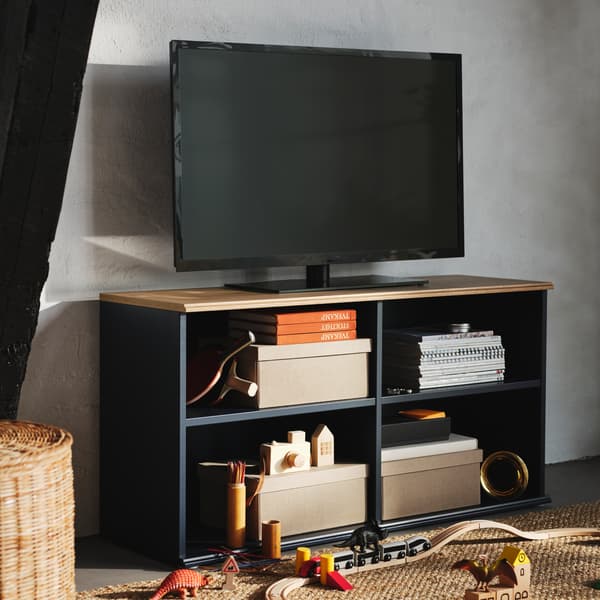 Kasser, bøger og legetøj ligger på de åbne hylder i et sort/blåt SKRUVBY tv-bord, hvorpå der står et sort tv.