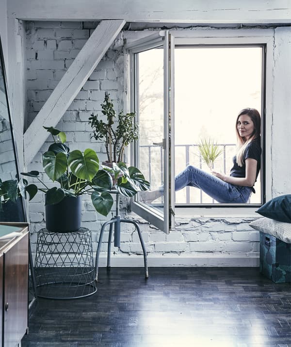 Katarzyna assise dehors sur l’appui de fenêtre d’une pièce avec des murs en briques peints en blanc et des plantes.