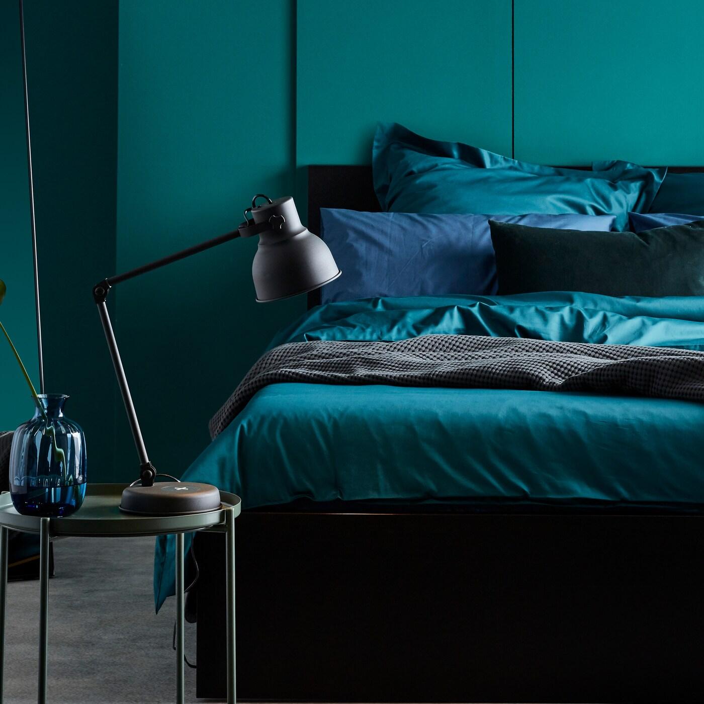 Katil MALM dengan tekstil katil termasuk linen katil berwarna hijau gelap terletak bersandar pada dinding di dalam bilik tidur.