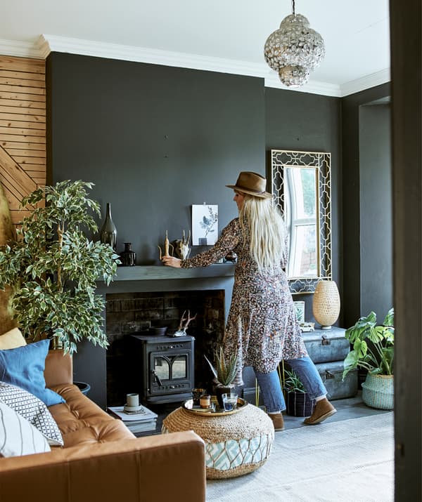 Katty arranging ornaments above a fireplace in a living room with dark walls and a brown leather sofa.