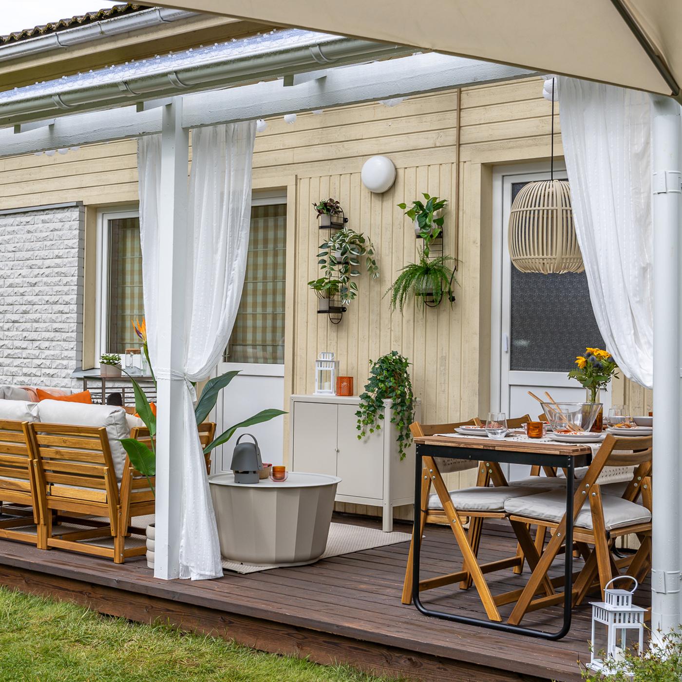Katusega terrassil on suur modulaarne diivan vasakul ja söögilaud koos toolidega paremal. Terrassi nurkades ripuvad valged kardinad. 