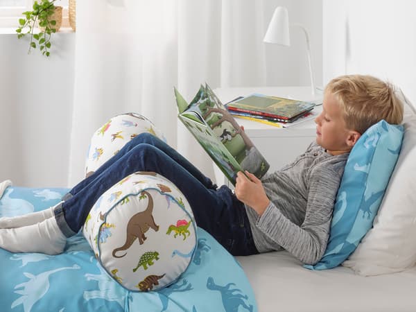 Kék JÄTTELIK ágyneművel behúzott ágyon olvasó fiút látunk, amint a lábát egy JÄTTELIK párnán nyugtatja A háttérben fehér függöny és fehér FUBBLA lámpa látszik, a lámpa egy MALM fiókos szekrényen áll.