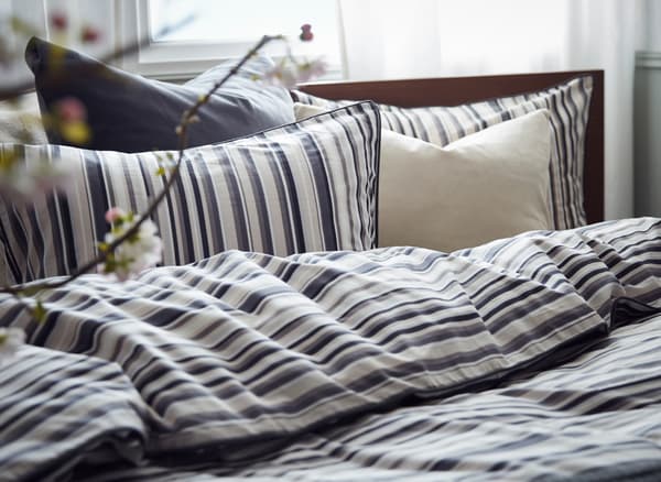 Kepala katil dengan linen katil kapas RANDGRÄS dengan bantal dan sarung kuilt berwarna putih dan kelabu, sesetengahnya berjalur.