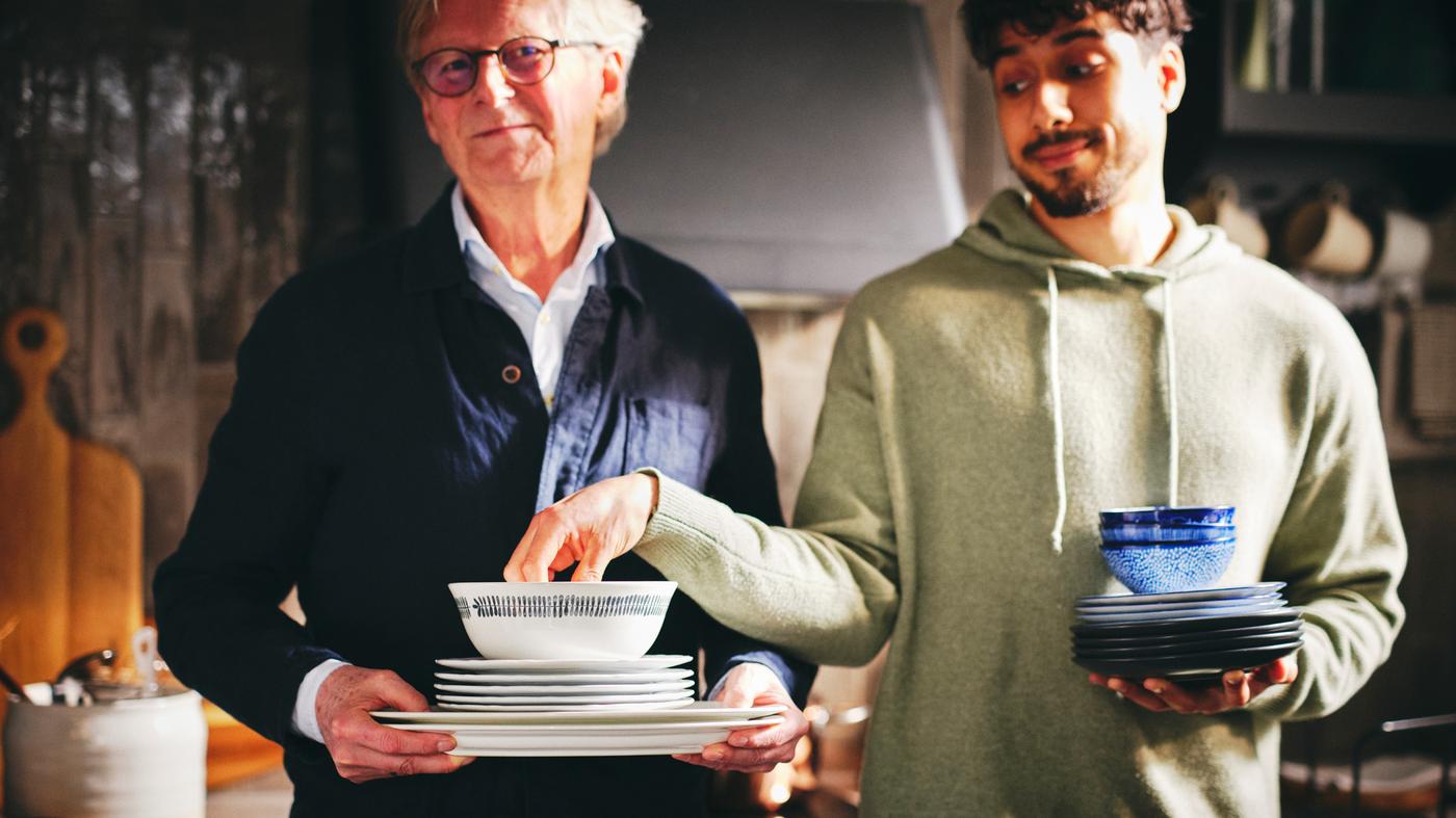 Két embert látunk, az egyikük egymásra tett fehér UPPLAGA tányérokat és tálakat, valamint egy fehér/mintás FRIKOSTIG tálat tart a kezében.