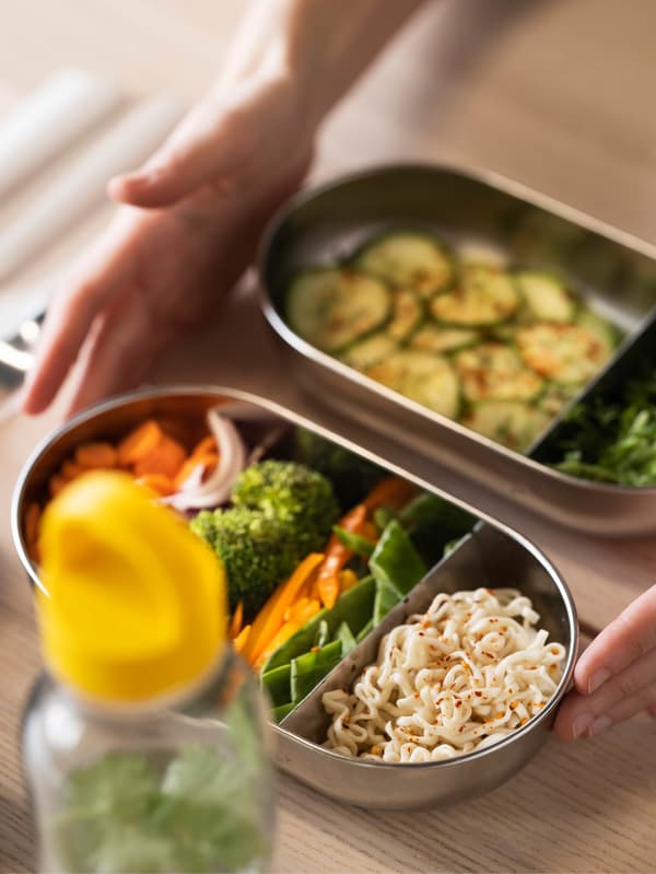 Két kezet látunk, amint egy HALVVARM ételtároló tetővel és elosztóval éppen az asztalra tesz. Az ételtárolóban zöldségek és tészta van. A háttérben egy másik ételtárolóban szeletekre vágott cukkinit, előtte pedig egy FORMSKÖN kulacsot látunk sárga fedővel.