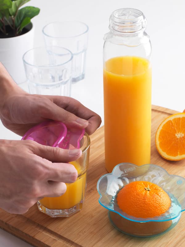 Két kezet látunk, amint pink szilikon ÖVERMÄTT ételfedőt tesz egy üveg narancslére. Mellette egy félbevágott narancs, rajta kék ételfedő, valamint egy üveg gyümölcslé, a tetején átlátszó ételfedővel.