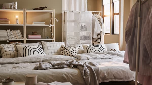 Két LYCKSELE LÖVÅS kinyitható kanapé szürke ágyneművel és mintás párnákkal, fehér-nyárfa polcok és fehér ruhatartók.