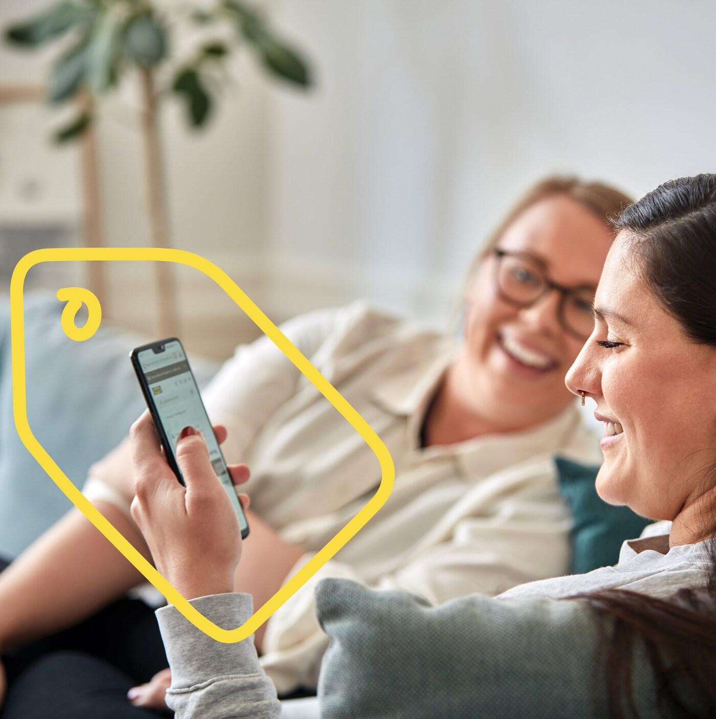Két nő ül egy kanapén, és nevetgélnek egy beszélgetés közepén. Az egyikük egy telefont néz, amely köré egy árcédula sárga képe van festve.