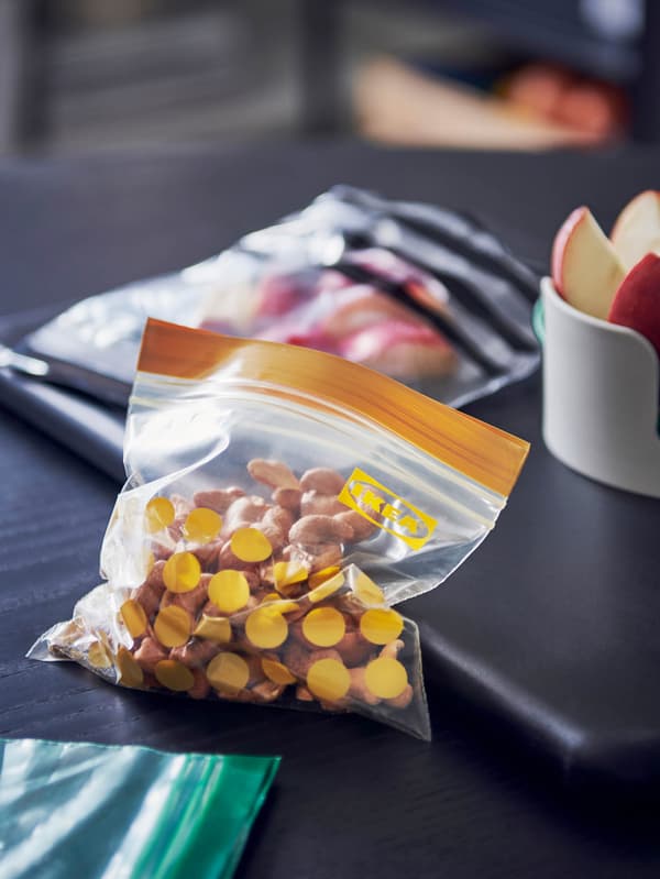 Két visszazárható ISTAD zacskó hever egy sötét asztallapon, az egyikbe diót, a másikba pedig szeletelt almát csomagoltak.