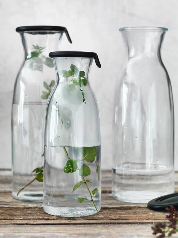 Két vízzel és fűszernövényekkel teli VARDAGEN kancsó egy faasztalon világosszürke fal előtt. 