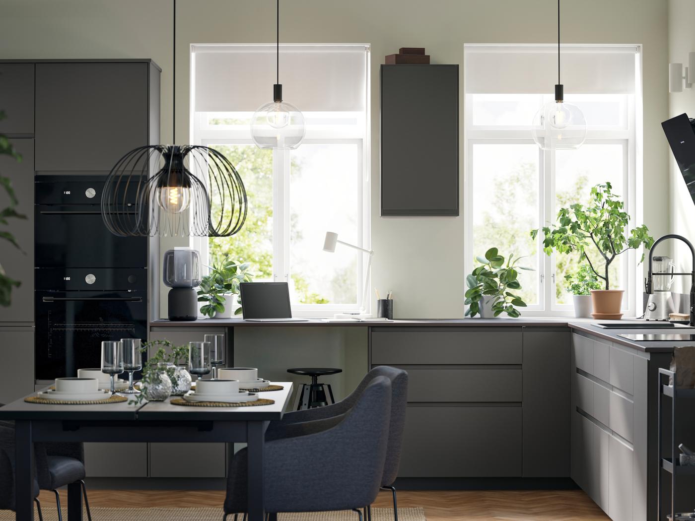 Keuken met vlakke, donkergrijze VOXTORP fronten heeft twee grote ramen, een eettafel, een hanglamp en zwarte toestellen.