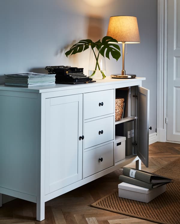 خزانة جانبية HEMNES مع مصباح والعديد من القطع الفنية في الأعلى. يكشف الباب الموارب عن صناديق ومجلدات مخزنة بداخلها.