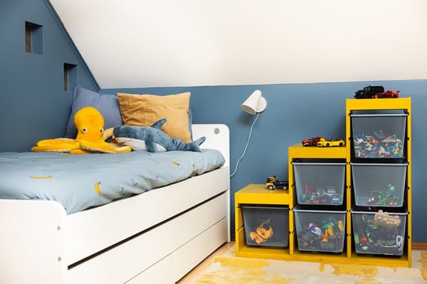Kids’ room with a white bed, pillows, and neatly organized toy storage boxes; a yellow octopus soft toy on the bed.