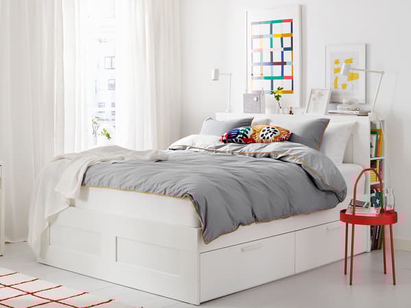 収納付き白いベッドにグレーの寝具を合わせた機能的な寝室。赤いサイドテーブルがアクセント。