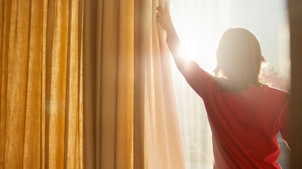 Kind in rood T-shirt trekt gordijnen in geel en wit open, zodat het zonlicht naar binnen stroomt.