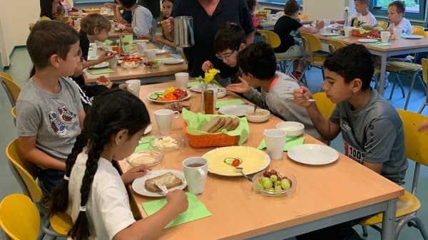 Kinder sitzen an einem Tisch in einer Schulkantine und essen gemeinsam ein Frühstück. Auf dem Tisch stehen Teller mit Brot, Käse, Gurken und Paprika sowie Tassen und Schalen mit Müsli. Eine erwachsene Person schenkt aus einer Kanne Tee oder Kaffee ein. Im Hintergrund sind weitere Kinder an anderen Tischen zu sehen.