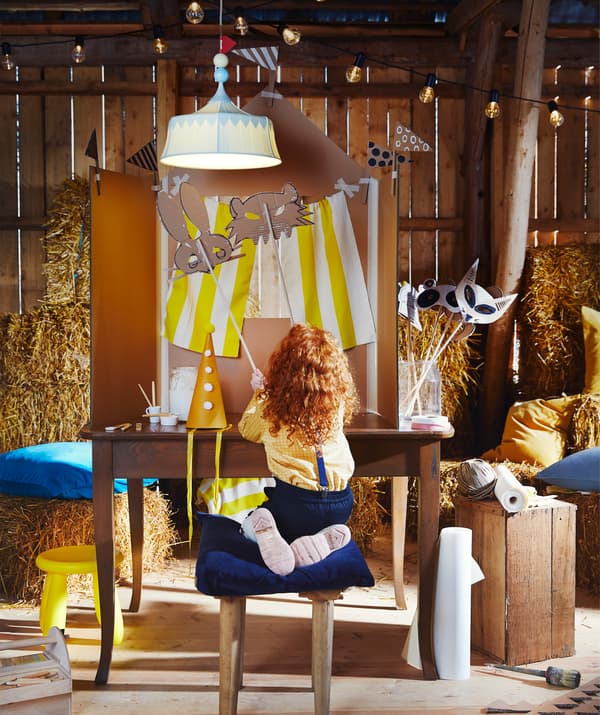 Kisgyerek egy kartonokból épült színpadon álarcokkal játszik a halványzöld mennyezeti lámpa alatt.