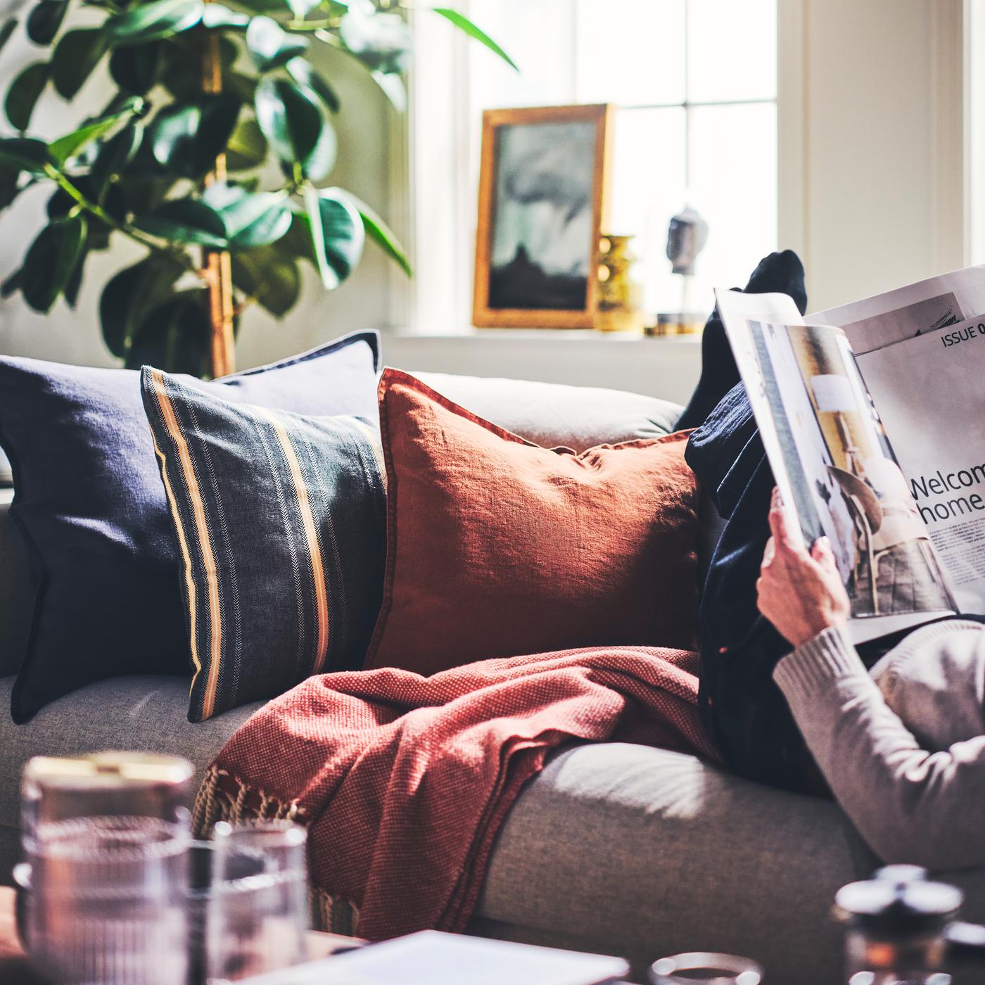 Kissen in einem STORTIMJAN Kissenbezug und in DYTÅG Kissenbezügen liegen auf einem Sofa. Daneben sitzt eine Person, die eine Zeitschrift liest.