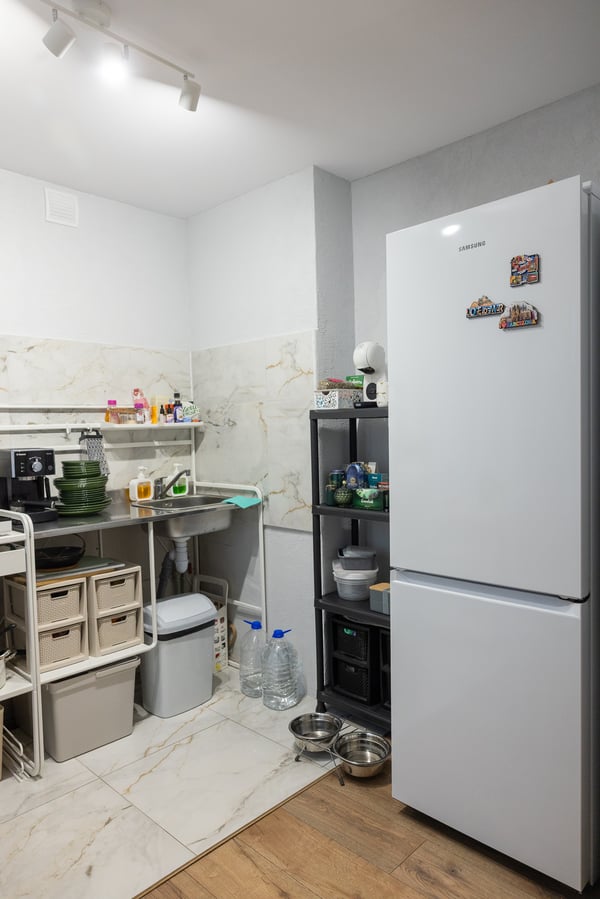 Kitchen area with a refrigerator and open shelving displaying boxes, jars and small appliances; a narrow walkway between the units.