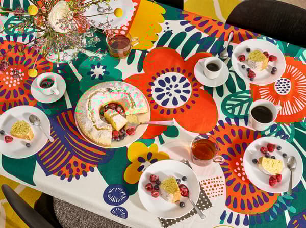 Kitchen with an overview of the kitchen table that has a flower-patterned tablecloth, plates, glasses, mugs and a cake on it.