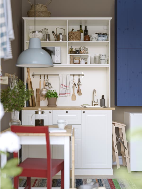 Kitchenette ÖNNERUP en blanc flanquée d’éléments IVAR peints en bleu. À l’avant-plan, petite table avec une chaise rouge.