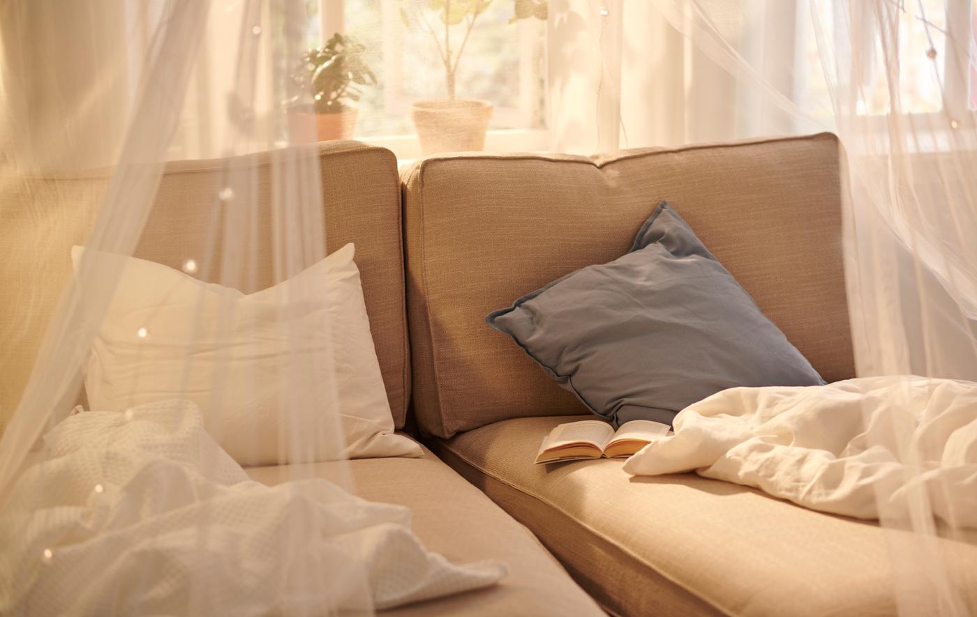 เก้าอี้ยาว KIVIK/ชีวิค สีเบจสองตัววางคู่กันริมหน้าต่างที่มีแสงแดดส่อง มีผ้าคลุมเตียงและหนังสือวางอยู่ มีมุ้ง SOLIG/ซูลลิก คลุมอยู่ด้านบน