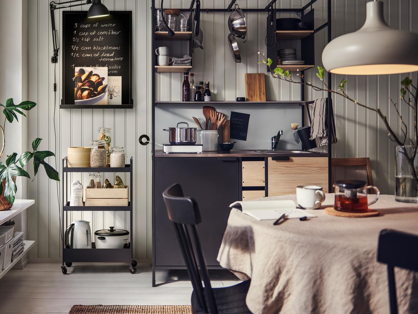 Kjøkken med ÄSPINGE minikjøkken med diverse ting i et svart NISSAFORS trillebord og et kjøkkenbord med stoler.