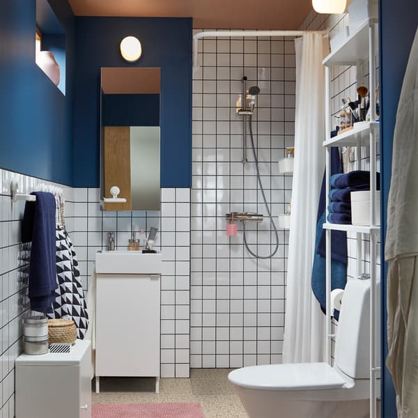 Kleine badkamer met douche, witte badkamermeubels, witte tegeld, donkerblauwe muren, roze badmat, spiegelkast en blauwe handdoeken.
