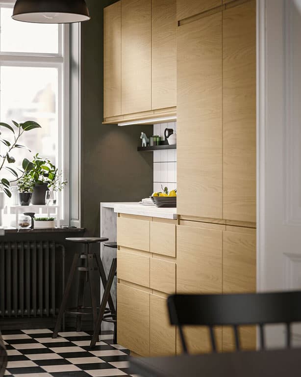 kleine IKEA Küchenlösung mit hölzernen Fronten. am Fensterbrett sind Dekopflanzen zu sehen. Unscharf im Vordergrund ein schwarzer Esstisch mit passendem schwarzen Hol Stuhl