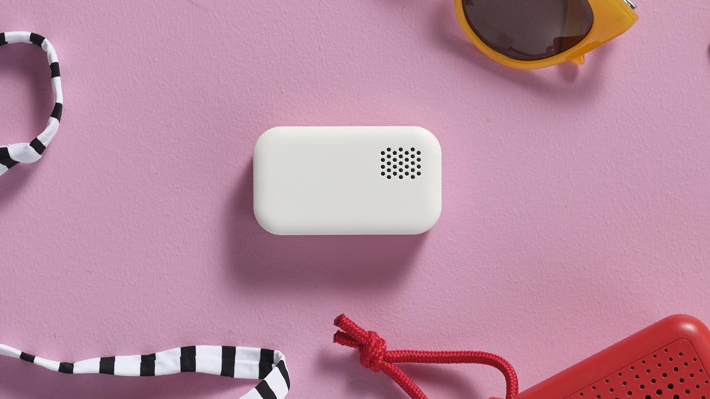 Kleiner weißer Lautsprecher liegt auf einer pinkfarbenen Oberfläche, umgeben von Sonnenbrille und bunten Bändern.

