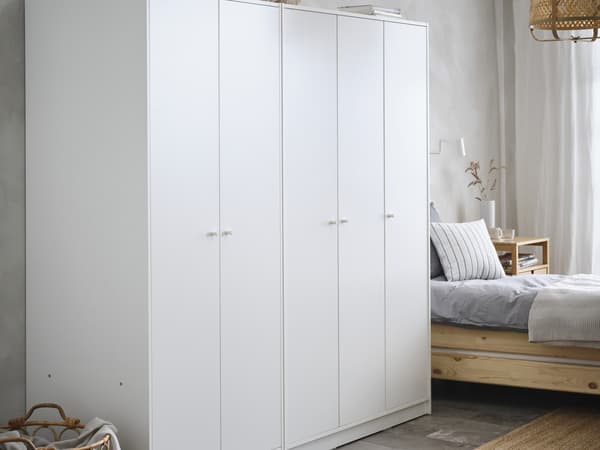 KLEPPSTAD wardrobe in white with hinged doors, in a bedroom against a grey cement screed wall and next to a natural wood bedframe with grey sheets. 