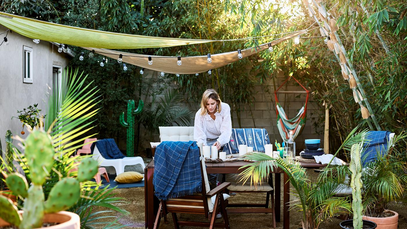 كلوي تقوم بترتيب الطاولة في الخارج في حديقتها، مع أثاث خشبي ونباتات وأشجار ومظلات مع سلاسل إضاءة.