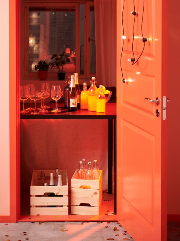 ประตูที่ตกแต่งสวยงามเปิดอยู่ มีโต๊ะเครื่องดื่มวางอยู่ตรงห้องโถง ขวดสำรองวางอยู่ในกล่องลัง KNAGGLIG/คนักกลิก ใต้โต๊ะ