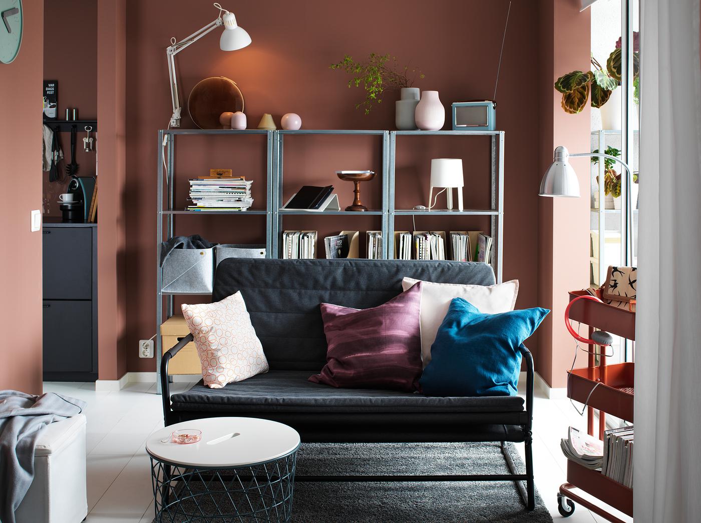 كنبة صغيرة رمادي داكن-أسود في غرفة جلوس مع جدران أحمر داكن ورفوف على الحائط الخلفي.