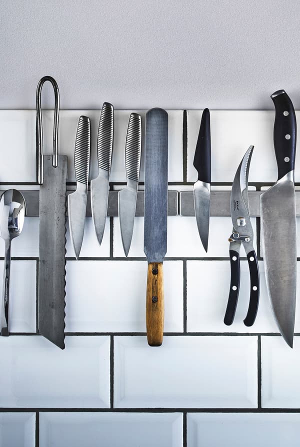 Knives and cooking utensils stored on a magnetic bar that is mounted on a white-tiled wall in the kitchen.