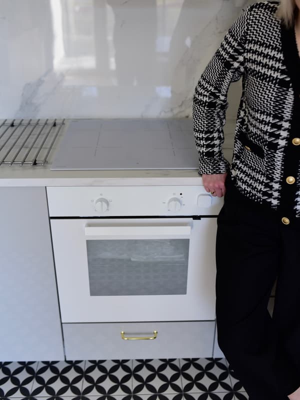 Kobieta oparta o szarą kuchnię, stojaca obok płyty i piekarnika w kolorze białym.